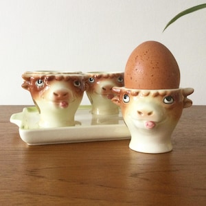 May include: A set of three ceramic egg cups shaped like brown and cream cow heads with small horns and tongues sticking out. One cup holds a brown speckled egg. The cups are arranged on a rectangular cream-colored ceramic tray with handles.