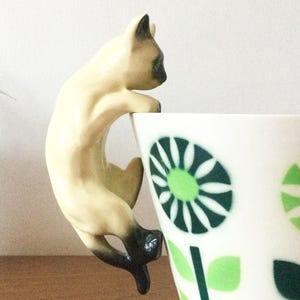 May include: A ceramic Siamese cat figurine with cream-coloured fur and dark brown points, designed to hang over the edge of a white teacup decorated with a repeating pattern of green and dark green stylised flower blossoms.