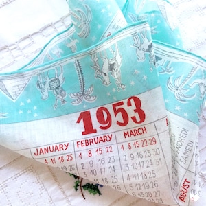May include: A vintage 1953 calendar tea towel featuring a light blue background with a repeating pattern of grey line-drawn palm trees and horses. The calendar displays the months of January, February, and March in red and black text.