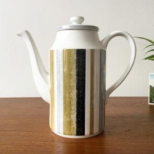 May include: A white ceramic coffee pot featuring a cylindrical body decorated with vertical stripes in shades of gold, black, and grey. The pot includes a matching lid with a small knob handle, a curved spout, and a large side handle for pouring.