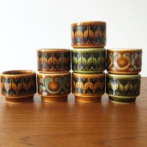May include: A set of seven ceramic egg cups arranged in a stack. The cups feature repeating geometric patterns in shades of brown, olive green, and black. Each cup has a cylindrical shape with a slightly flared rim and a stepped base design.