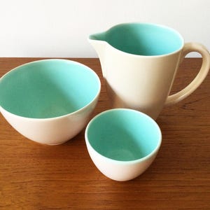 May include: A ceramic pitcher, a large bowl, and a small bowl, each featuring a smooth off-white exterior and a vibrant turquoise interior. The pitcher has a curved handle and a pouring spout, while the two bowls have rounded, deep basins.
