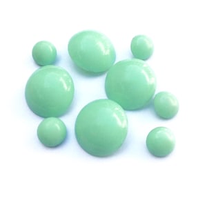 Könnte beinhalten: Eine Ansammlung von acht glatten, runden, mintgrünen Cabochons in verschiedenen Größen. Jedes Stück weist eine polierte, gewölbte Oberfläche mit glänzendem Finish auf und ist in einer Gruppe angeordnet. Die Objekte sind einfarbig.