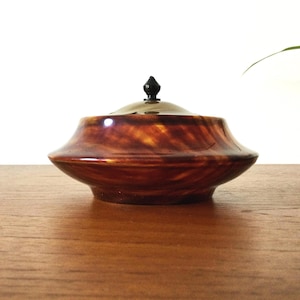 May include: A polished, round wooden bowl with a glossy, swirling amber and dark brown grain pattern. The bowl features a matching domed lid topped with a small, pointed black finial handle, creating a smooth, symmetrical, and low-profile silhouette.