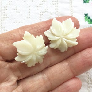以下が含まれることがあります： オフホワイトの彫刻が施された花形のボタンまたは装飾用アクセントのペア。各ピースは、滑らかで光沢のある仕上げの多層的な花びらのデザインが特徴で、繊細な彫刻が施された様式化された花や葉の集まりを思わせる形状をしています。