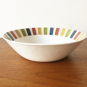 Peut inclure: Un bol en céramique blanche avec un extérieur texturé et un large bord orné d'un motif répétitif de rayures verticales dans des tons sourds de vert olive, gris anthracite et orange terre cuite, séparées par de fines lignes noires.