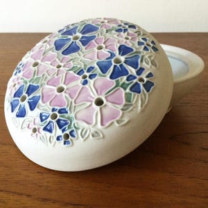 May include: A round, off-white ceramic flower frog or lidded box featuring a raised, hand-painted floral pattern in shades of blue, pink and green. The lid is domed and includes several circular perforations distributed among the flower petals.