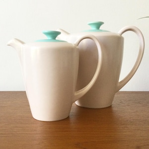 May include: Two off-white ceramic coffee pots with smooth, tapered bodies and curved handles. Each pot features a small, circular, light turquoise lid with a central knob. The pots are positioned side-by-side, showcasing a clean, simple design.
