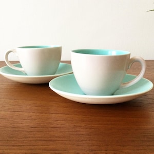 May include: Two ceramic tea cups and matching saucers featuring a matte white exterior and a smooth, light turquoise interior. Each cup has a curved handle and sits centred on a round saucer that shares the same two-tone colour scheme.