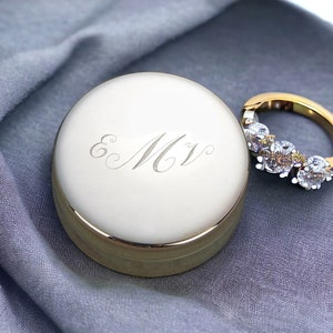 May include: A gold ring with three large, clear gemstones and smaller brown and yellow stones. Beside the ring is a round, gold-colored box with the engraved initials "E M V" on the lid. The items rest on a gray fabric.