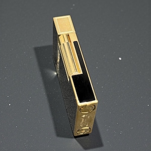 May include: A gold-colored lighter with a rectangular shape and black accents. The lighter has a shiny finish and a small rectangular top. The bottom of the lighter has two circular features and some engraved text.