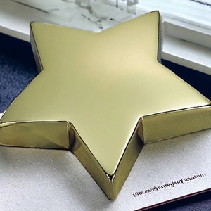 May include: A gold-colored star-shaped paperweight. The star is sitting on a white surface with a brown border.