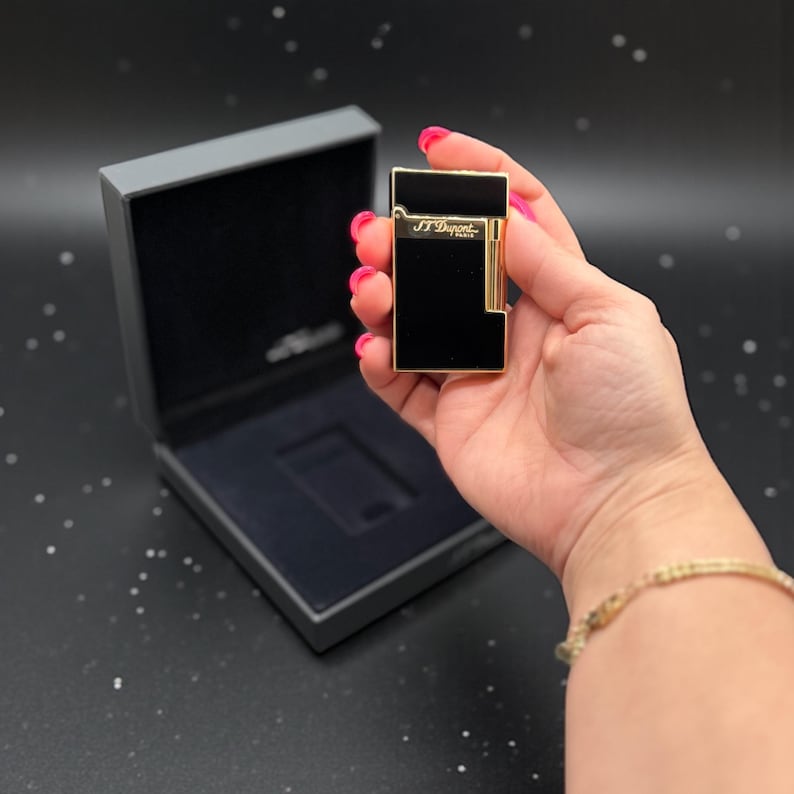 May include: A black and gold lighter with the text "S.T. Dupont Paris" is held in a hand. The lighter is rectangular with gold trim and a black surface. The lighter is in front of a black box.