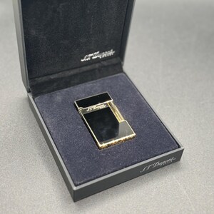 May include: A black and gold lighter in a black presentation box. The lighter has a rectangular shape with gold accents and the brand name "S.T. Dupont" is visible on the lighter and the box.