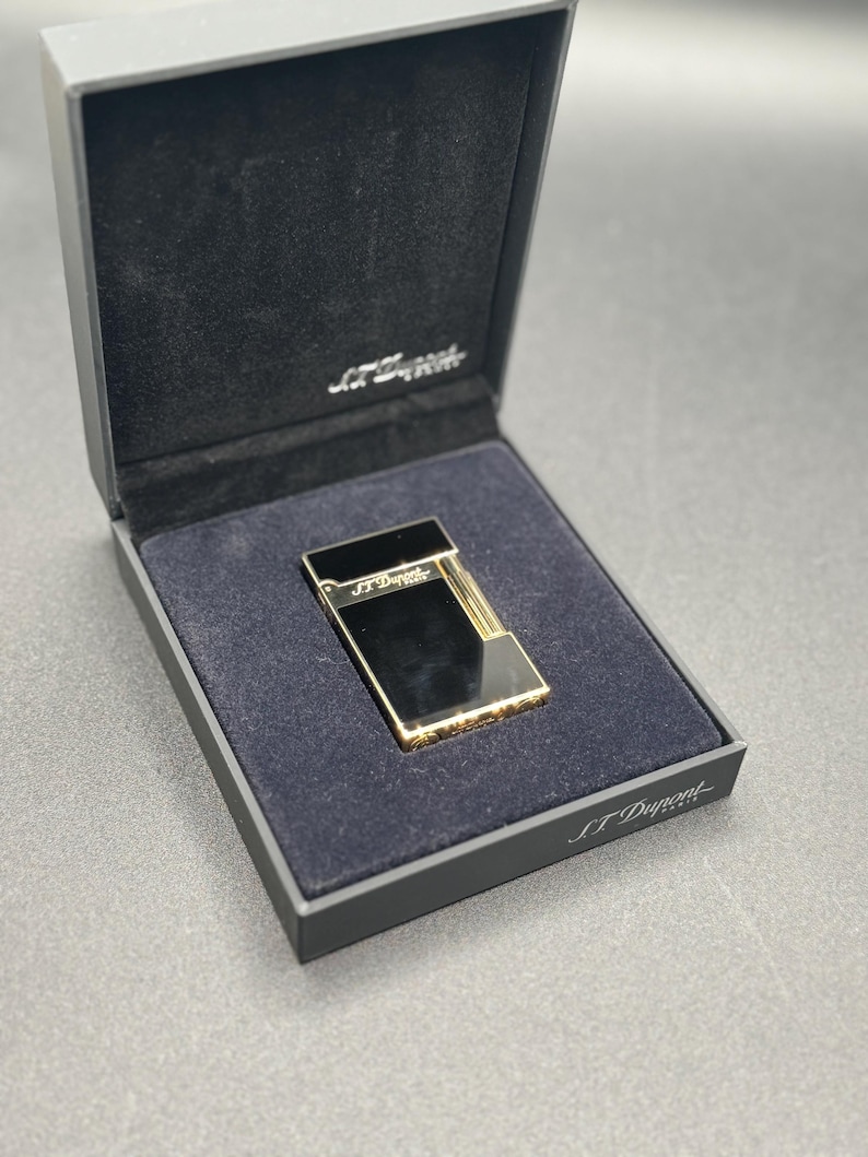 May include: A black and gold lighter in a black presentation box. The lighter has a rectangular shape with gold accents and the brand name "S.T. Dupont" is visible on the lighter and the box.