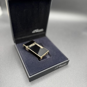 May include: A black and gold lighter in a black box with a blue interior. The lighter has a rectangular shape with gold trim and a black surface. The box is open, revealing the lighter inside. The brand name is visible on the box.