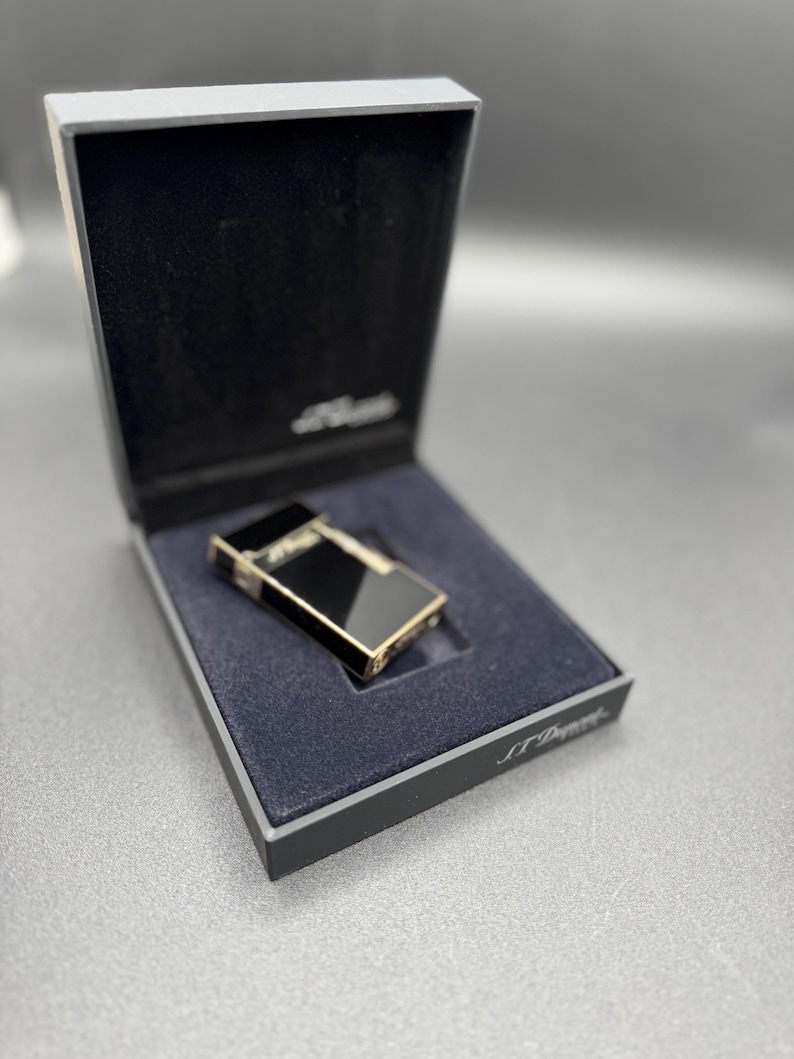 May include: A black and gold lighter in a black box with a blue interior. The lighter has a rectangular shape with gold trim and a black surface. The box is open, revealing the lighter inside. The brand name is visible on the box.