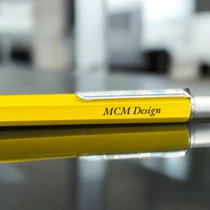 Könnte beinhalten: Ein gelber Stift mit einer silbernen Klammer und dem Aufdruck "MCM Design" auf der Seite.