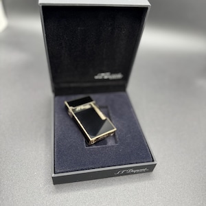 May include: A black and gold lighter in a black box. The lighter has a rectangular shape with a shiny black surface and gold trim. The box is open, revealing a dark blue interior and the brand name "S.T. Dupont".