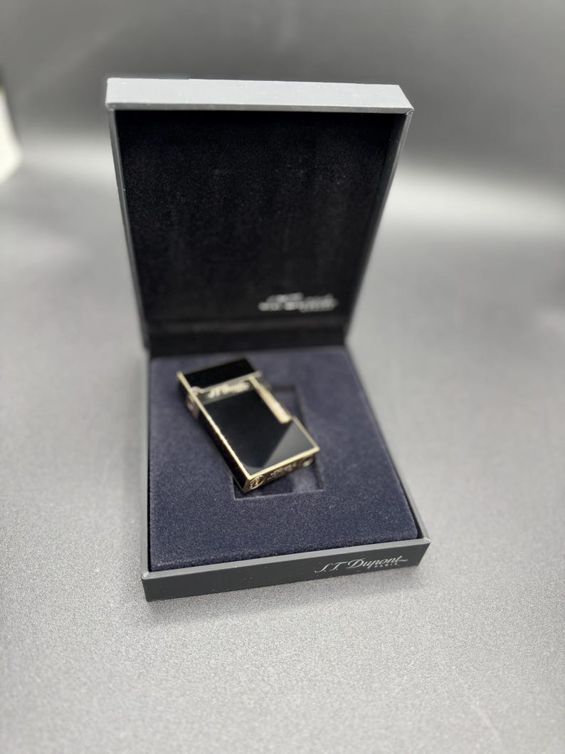 May include: A black and gold lighter in a black box. The lighter has a rectangular shape with a shiny black surface and gold trim. The box is open, revealing a dark blue interior and the brand name "S.T. Dupont".