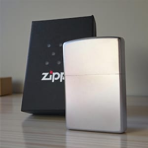 May include: A silver-colored Zippo lighter with a rectangular shape, next to a black box with the Zippo logo. The lighter has a brushed metal finish. The box has the Zippo logo and a series of dots.