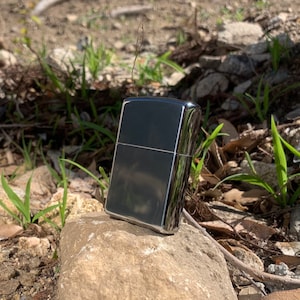 May include: A silver lighter with a black finish. The lighter is sitting on a rock with green plants in the background.