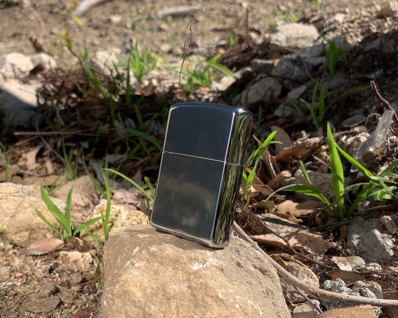 May include: A silver lighter with a black finish. The lighter is sitting on a rock with green plants in the background.