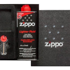 May include: A black box with a red and white Zippo lighter fluid bottle and a silver lighter inside. The bottle is labeled "Zippo Lighter Fluid" and has a red cap. The box has a black background with the word "Zippo" in white letters.