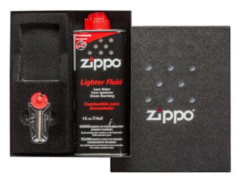 May include: A black box with a red and white Zippo lighter fluid bottle and a silver lighter inside. The bottle is labeled "Zippo Lighter Fluid" and has a red cap. The box has a black background with the word "Zippo" in white letters.