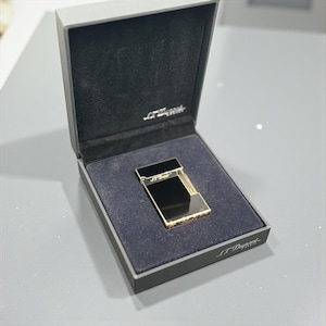 May include: A gold and black lighter in a black box with the brand name "S.T. Dupont" printed on the box. The lighter has a rectangular shape with a shiny gold finish and a black top.
