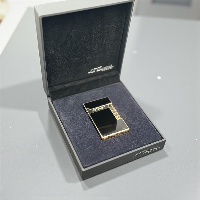 May include: A gold and black lighter in a black box with the brand name "S.T. Dupont" printed on the box. The lighter has a rectangular shape with a shiny gold finish and a black top.