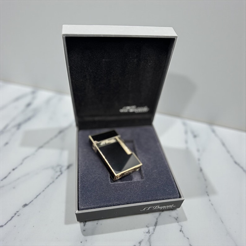 May include: A black and gold lighter in a presentation box. The lighter has a rectangular shape with a black top and gold accents. The box is white with a black interior and the brand name "S.T. Dupont" is visible.