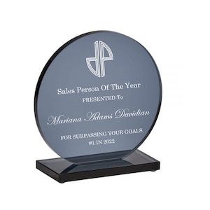 May include: A blue glass award with a black base. The award is inscribed with the text "Sales Person Of The Year" and "Presented To Mariana Adams Davidian" and "For Surpassing Your Goals #1 In 2022".