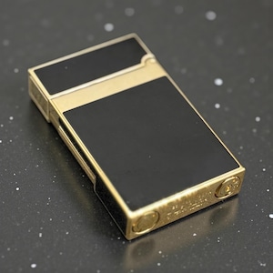 May include: A rectangular lighter with a gold-colored frame and black panels. The lighter has a classic design with a hinged lid and a textured surface. The lighter is a stylish accessory.