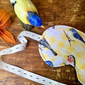 May include: A white sleep mask with a yellow background and a pattern of colorful birds. The mask has an elastic strap with adjustable buttons.