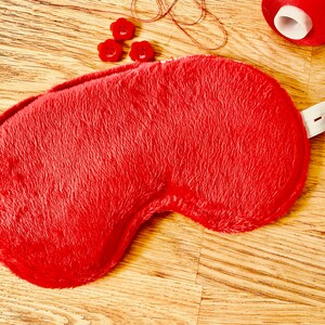 May include: A red fuzzy sleep mask with a white elastic strap. The mask is lying on a wooden surface.