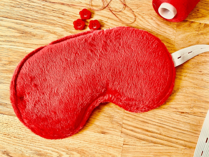 May include: A red fuzzy sleep mask with a white elastic strap. The mask is lying on a wooden surface.