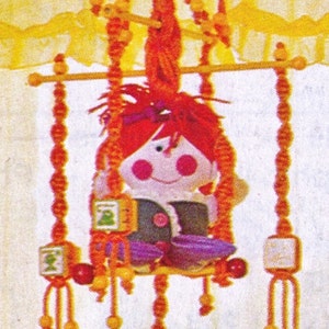 May include: A red and yellow crocheted mobile with a hanging doll. The doll has red hair, a pink face, and black eyes. The mobile has small wooden blocks with different colored designs.