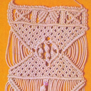 Peut inclure: Suspension murale en macramé de couleur crème avec des motifs géométriques. Le design présente des formes de losanges et de triangles, avec des détails noués et des franges. Le fond est de couleur orange uni.