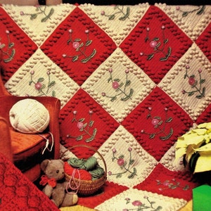 Könnte beinhalten: Eine rot-cremefarbene Häkeldecke mit Rautenmuster und Blumenstickerei. Die Decke liegt auf einem roten Sessel mit einer Wollknäuel und einem Teddybären.