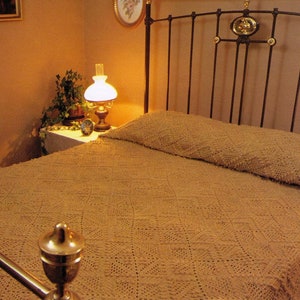 May include: A vintage iron bed frame with a crocheted bedspread and a white lamp on a side table. The bedspread is a light brown color and the lamp has a white shade.