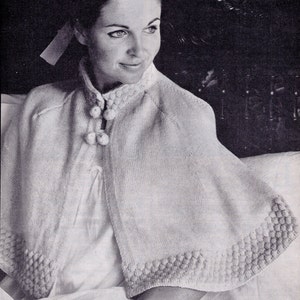 May include: A white knitted cape with a button fastening and a textured border. The cape is being worn by a woman with dark hair and a white blouse.