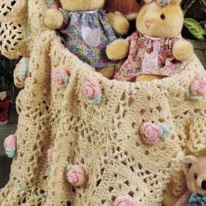 May include: A crocheted baby blanket with a delicate floral design. The blanket is cream coloured with pink and green flowers. Three plush bunny toys are sitting on the blanket.