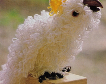 May include: A white yarn cockatoo with a yellow crest sits on a wooden platform. The cockatoo has a wooden beak and black yarn feet.