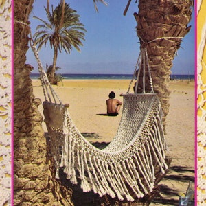 May include: A white hammock with fringe hangs between two palm trees on a sandy beach. A person is sitting in the hammock.