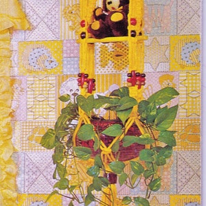 May include: A yellow macrame plant hanger with a brown teddy bear sitting in the centre. The hanger is decorated with red beads and has a fringe at the top. The hanger is hanging on a wall with a yellow and pink patchwork design.