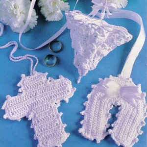 May include: Three white crocheted items: a cross, a horseshoe shape, and a bell-shaped object. The items are on a blue background with two silver rings.
