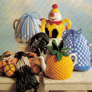 Könnte beinhalten: Eine Sammlung gestrickter Teekannenwärmer in verschiedenen Farben und Mustern, darunter ein blau-weiß kariertes Design, ein gelbes und grünes Ananas-Design, ein blau-weiß gestreiftes Design und ein schwarz-grünes Design. Die Teekannenwärmer werden auf einer weißen Oberfläche präsentiert.