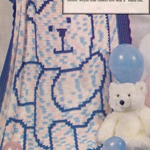 Op de afbeelding: Een gehaakt teddybeer-deken in roze, wit en blauwe ombre met een koninklijk blauwe omtrek. Het deken ligt op een lichtbruin oppervlak met een witte teddybeer en blauwe ballonnen.