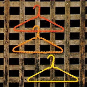 May include: Three wire hangers in different colors: red, orange, and yellow. The hangers are stacked on top of each other against a wooden background.